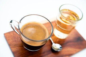 Crema of black coffee in a clear glass on a wooden tray, with a clear tea glass next to it selective focus, soft focus.           