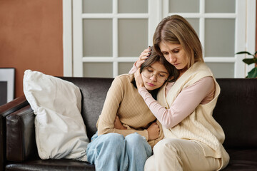 Mother sitting on sofa together with her daughter and embracing her, she comforting her during...
