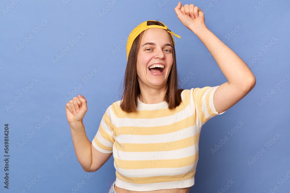 Wall mural Portrait of extremely happy joyful teenager girl wearing baseball cap and stripe T-shirt posing isolated over blue background, spending free time at party, dancing and smiling.