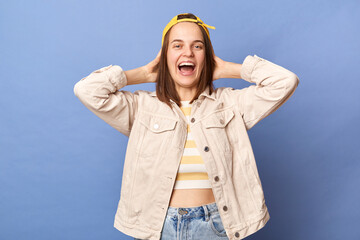Photo of extremely happy cheerful satisfied brown haired teenager girl in baseball cap and jacket, keeps hands behind her head, looking at camera and laughing, posing isolated over blue background.