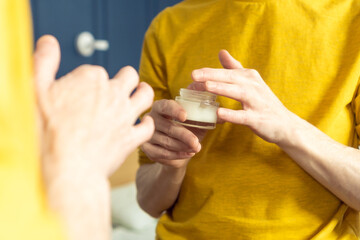 Hands of middle aged man holding jar of moisturizing cream. Crop male portrait in room mirror. Skincare morning routine, careful beauty treatment, home lifestyle. Facial care concept.