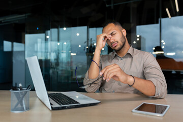 Tired and upset businessman inside office at work sitting at workplace with laptop, young african...