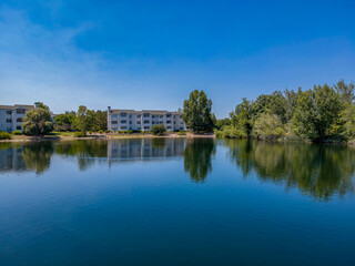 Fototapeta na wymiar Apartments with beautiful view of a reflective small lake in Boise Idaho. The lush green trees are reflected on the calm water surface with bright blue sky overhead on this sunny day.