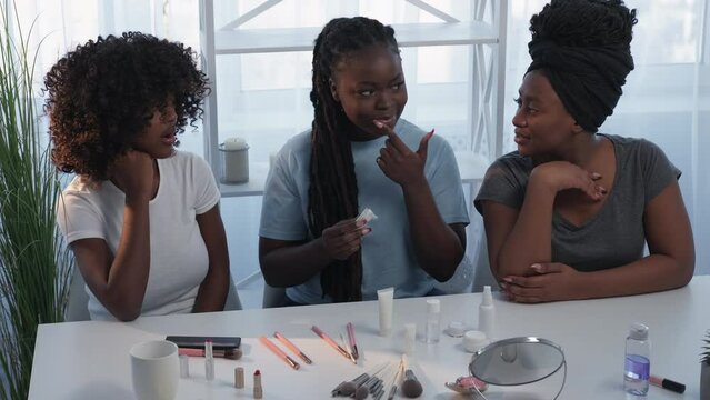 Beauty consultation. Female friends. New product. Inspired happy black women testing smelling decorative cosmetic sitting home desk in light room interior.