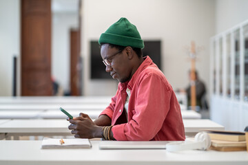 Smiling black guy using smartphone, sitting in library, happy young African male student scrolling on social media, chatting online during study break. Mobile phone addiction, academic procrastination