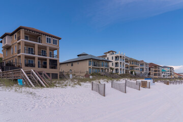 Sand dunes with slanted wood fences at the front of beach houses in Destin, Florida. There are footbridges access to the beach with white sand at the front.