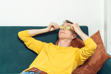 Adult man lying on sofa put patches from fresh cucumber slices on eyes in living room. Middle aged person reclining on pillow with mask for dark circles under eyes. Home relaxation, beauty treatment.