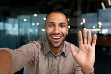 Smiling and cheerful businessman in office taking selfie photo on phone and talking on video call with colleagues and friends using smartphone, african american man waving at camera greeting gesture. - Powered by Adobe