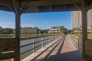 Views of a wooden boardwalk heading to residences under a gazebo in Destin, Florida. Views under the roof of a gazebo connected to a boardwalk over the lake heading to the apartments at the back.