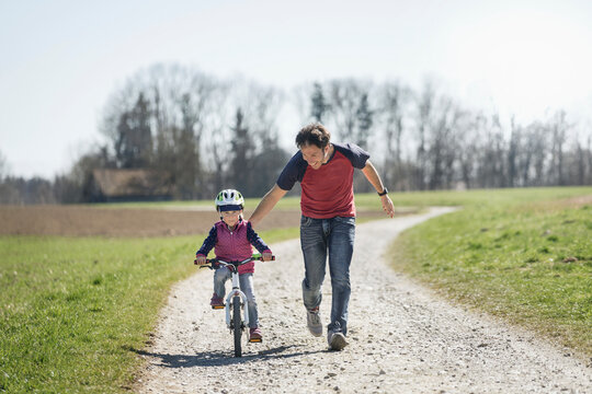 Father assisting daughter riding bicycle on footpath amidst field