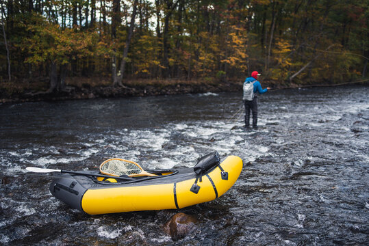 A fly-fisherman casting behind a packraft on a river in fall