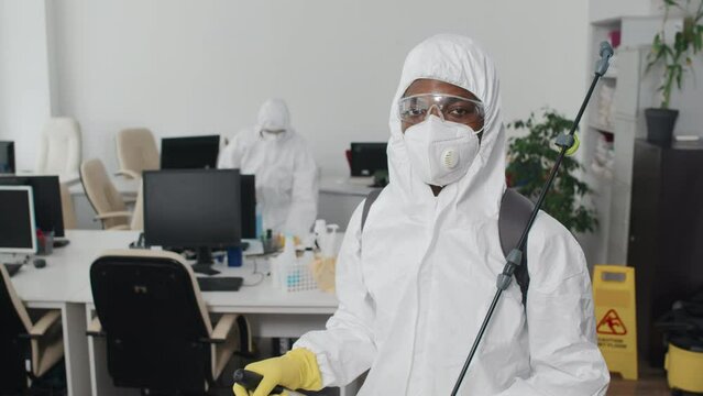 Selective focus portrait of unrecognizable Black man providing disinfection service wearing protective overall with eyewear and mask standing in office looking at camera