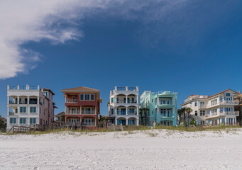 Fototapeta na wymiar Beach houses facade with balconies and roof decks facing the white sand shore in Destin, Florida. There are footbridges as beach access over the sand dunes at the front of the colorful beach houses.