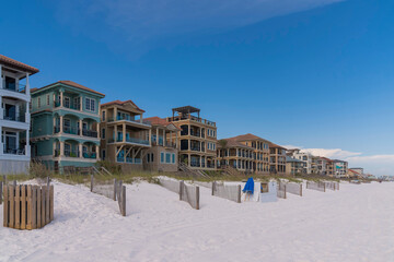 Side view of beach houses with fenced sand dunes and footbridge at the front in Destin, Florida. Row of multi-storey homes against the blue sky background.