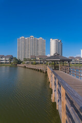 Obraz premium Vertical shot of wooden footbridge over Stewart Lake in Destin, Florida. Walkway with railings heading to the apartments and hotels with low-rise and high-rise structures against blue sky.