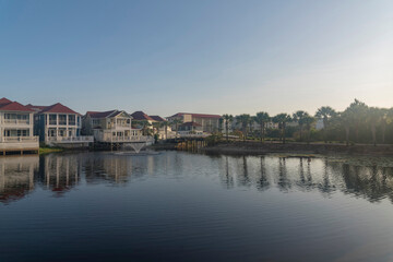 Fototapeta na wymiar Decorative lake with reflections of the waterfront houses in Destin, Florida. There is a lake at front with water fountain near houses with decks and balconies on the left and palm trees on the right.