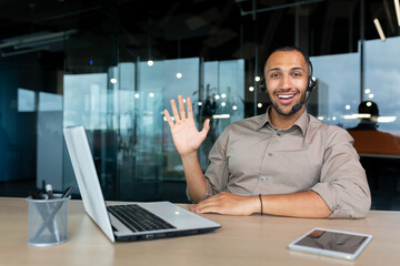 Portrait of tech support and online customer service worker, man with video call headset smiling and looking at camera, african american man in shirt working inside office using laptop.