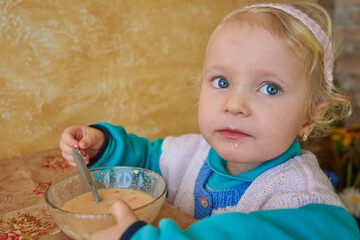 Naklejka premium little girl eats porridge,a beautiful child at the table with a bowl of porridge, beautiful blue eyes