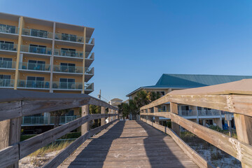 Wooden bridge with railings heading to the apartments or hotels with palm trees in Destin, Florida. Walkway over the sand dunes with grass near the multi-storey buildings against sky background.