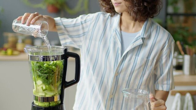 Tilt up shot of young woman adding glass of water to blender while preparing green smoothie in kitchen at home