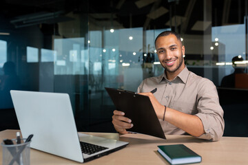 Portrait of smiling and satisfied businessman with achievement results, african american paper work smiling and looking at camera, young financier writing document inside office using laptop and pen.
