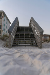 Open wooden stairs view from a white sand below at dusk in Destin, Florida. Wooden stairs to a footbridge over sand dunes near beach houses against dusk sky at the background.