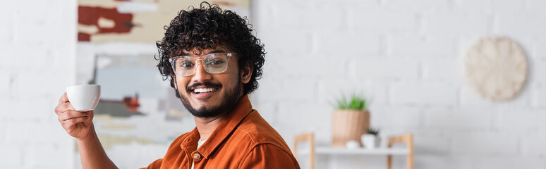 Smiling indian man in eyeglasses holding cup of coffee at home, banner.