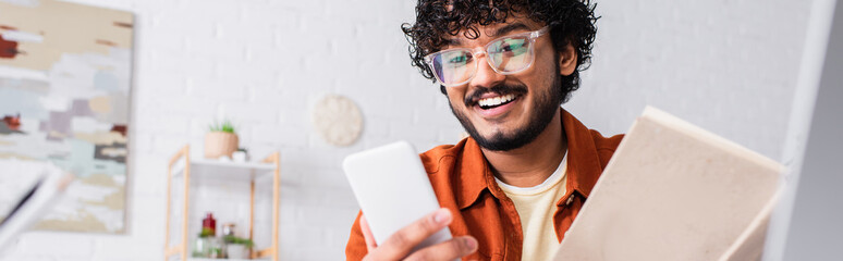 Cheerful indian copywriter in eyeglasses using smartphone and holding book at home, banner.