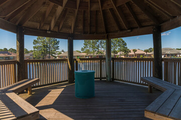 Wooden gazebo with seats and views of the lake and houses in Navarre, Florida. Gazebo on a lake with blue barrel trash bin with lid at the center.