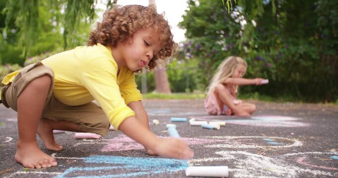 Little mixed race boy happily drawing chalk pictures on the tarmac walkway in a park