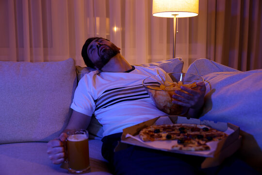 Man with chips, pizza and glass of beer sleeping on sofa at night. Bad habit