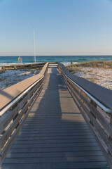 Fototapeta na wymiar Wooden pathway along with the grassy sand dunes heading to the beach at Destin, Florida. Footbridge with railings and views of blue ocean and skyline background.