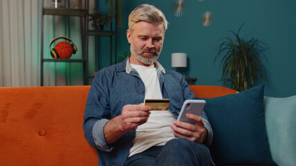 Middle-aged old man using credit bank card and smartphone while transferring money, purchases online shopping, order food delivery at home apartment indoors. Mature senior guy in evening room on couch