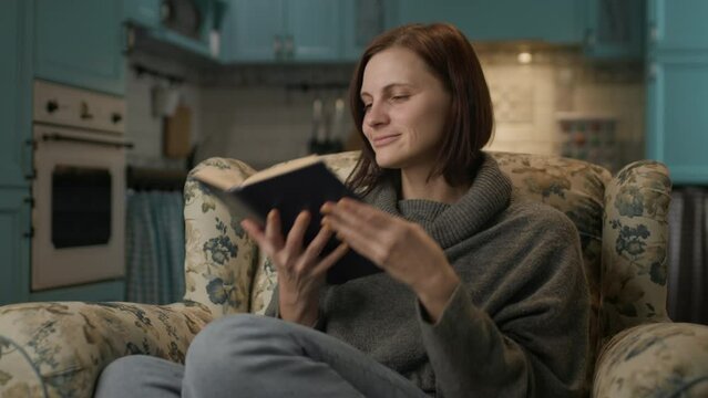 Woman Reading Paper Book. 30s female smelling book pages sitting on cozy couch at home.