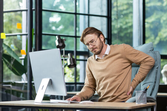 Tired businessman at workplace has severe back and lower back pain, young man working sitting at table with laptop and computer in middle of office.