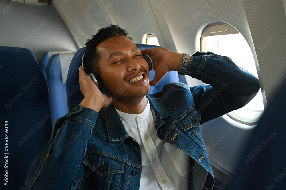 Wall mural happy asian man in wireless headphones listening to music and sitting comfortable seat in airplane c