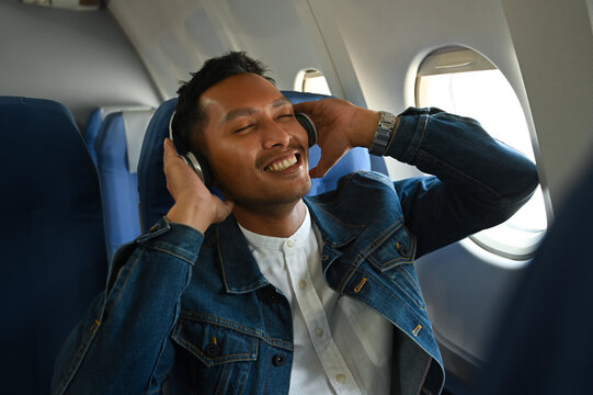 Happy Asian Man In Wireless Headphones Listening To Music And Sitting Comfortable Seat In Airplane Cabin