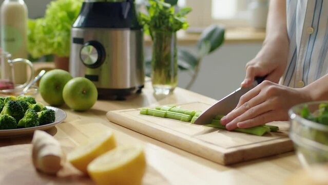 Close up shot of female hands cutting fresh celery stalks on wooden board at kitchen table while preparing healthy dinner at home