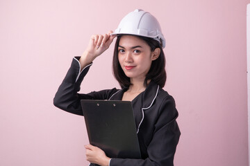 Beautiful young woman wearing white safety helmet and holding clipboard isolated on pink background
