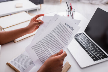 Cropped view of freelancer holding papers with copywriting lettering near book and laptop at home. - obrazy, fototapety, plakaty