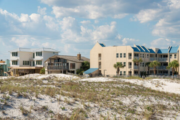 Sand dunes near the residential buildings against the puffy clouds in the sky at Destin, Florida. There is a fenced apartment with blue roofs and palm trees outdoors on the right.