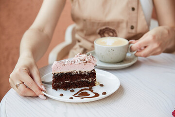 Woman holds fruit chocolate cake and cup of cappuccino on wooden table outside in the cafe terrace. Atmospheric coffee time, time for yourself. Heart shaped dessert decorations.