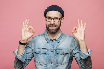 Cheerful bearded male in fashionable clothing, makes ok sign, being satisfied with something, isolated over pink studio background. Unshaven middle aged man gestures indoor, shows his approval