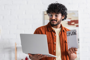 Indian copywriter holding notebook and laptop at home.