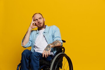 A man in a wheelchair problems with the musculoskeletal system looks at the camera thoughtfully with tattoos on his arms sits on a yellow studio background, health concept a person with disabilities