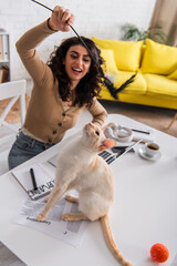 Blurred freelancer playing with oriental cat near documents and devices on table at home.