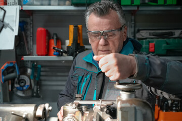 A man in safety glasses and a work uniform sets up a machine in his workshop. The grinding machine details in focus. In the background are various working tools on the metal shelving for workshop.