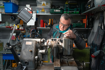 A man in safety glasses and work uniform sets up a grinding machine in his workshop. The grinding machine details in focus. In the background are various working tools on metal shelving for workshop.