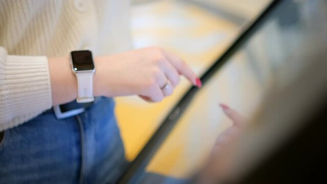 Woman using touchscreen terminal in mall. Detail view outstretched female hand touching modern big timetable or info screen in interior. Side view fingers swiping on display at out of focus background