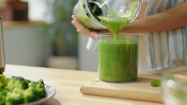 Close up shot of hands of woman pouring freshly made green smoothie from blender into glass